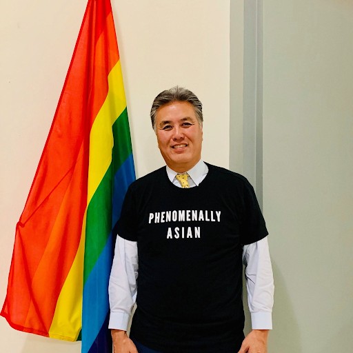 Rep. Mark Takano standing with a Pride flag wearing a Phenomenally Asian shirt